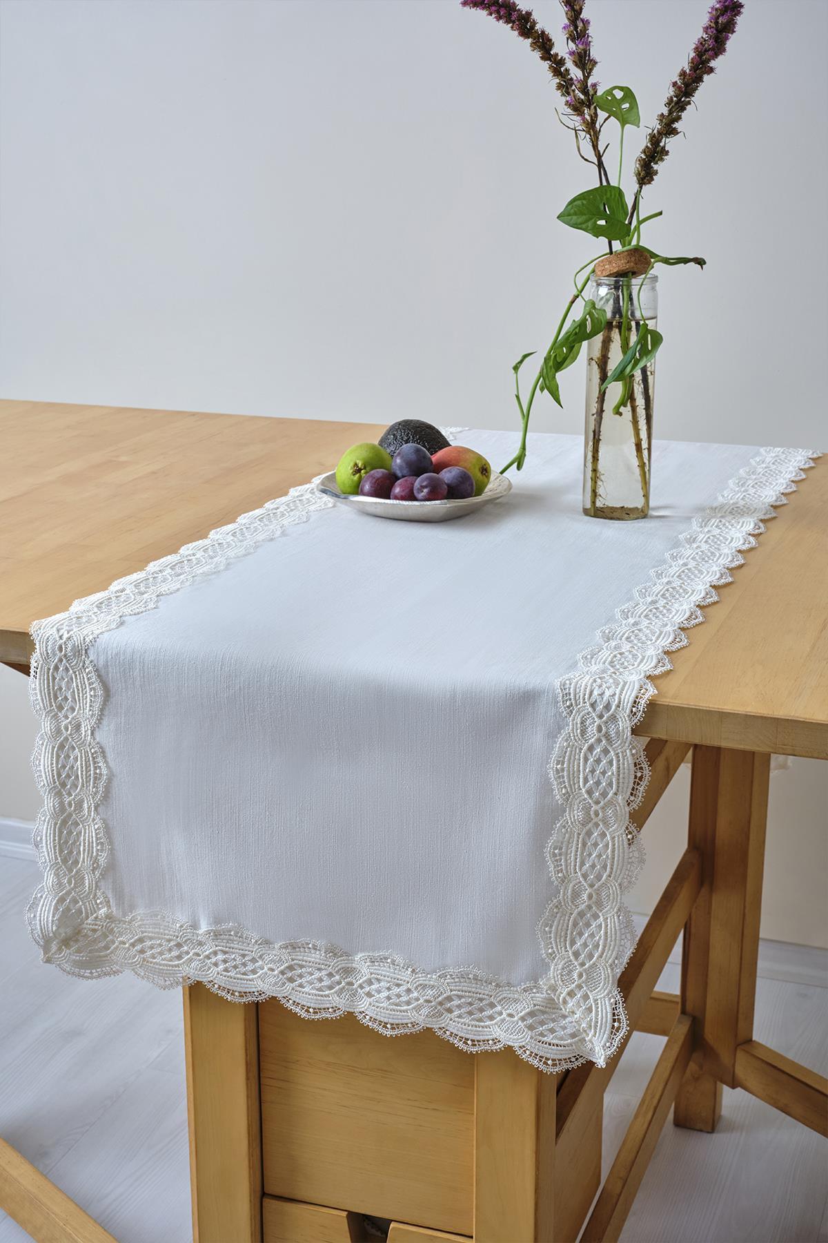 Chemin de table blanc avec détails en dentelle, accompagné d'une coupe de fruits et d'un vase en verre sur une table en bois