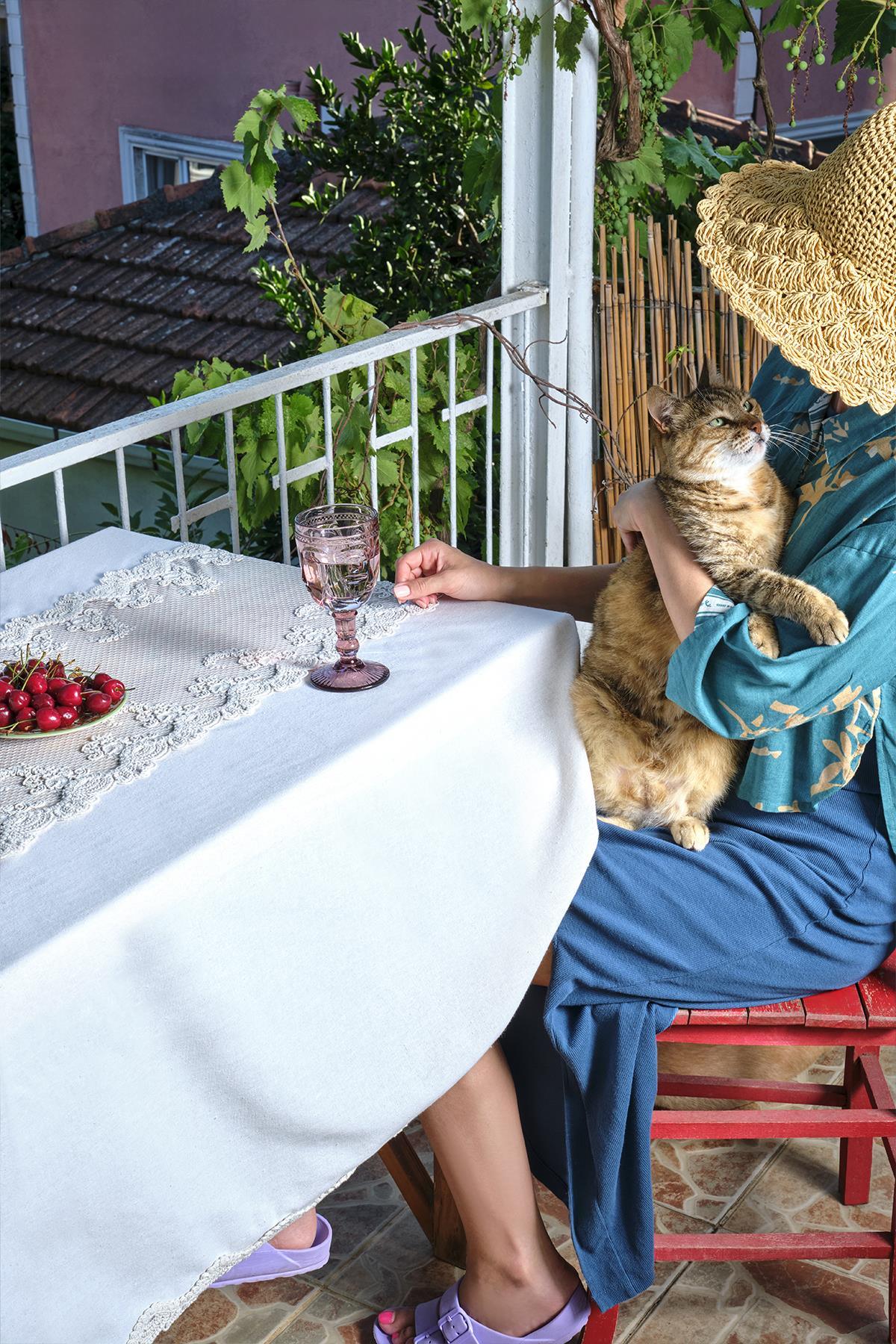 Nappe en dentelle sur une table de balcon, avec des cerises et un verre rose, accompagnée d'un chat.