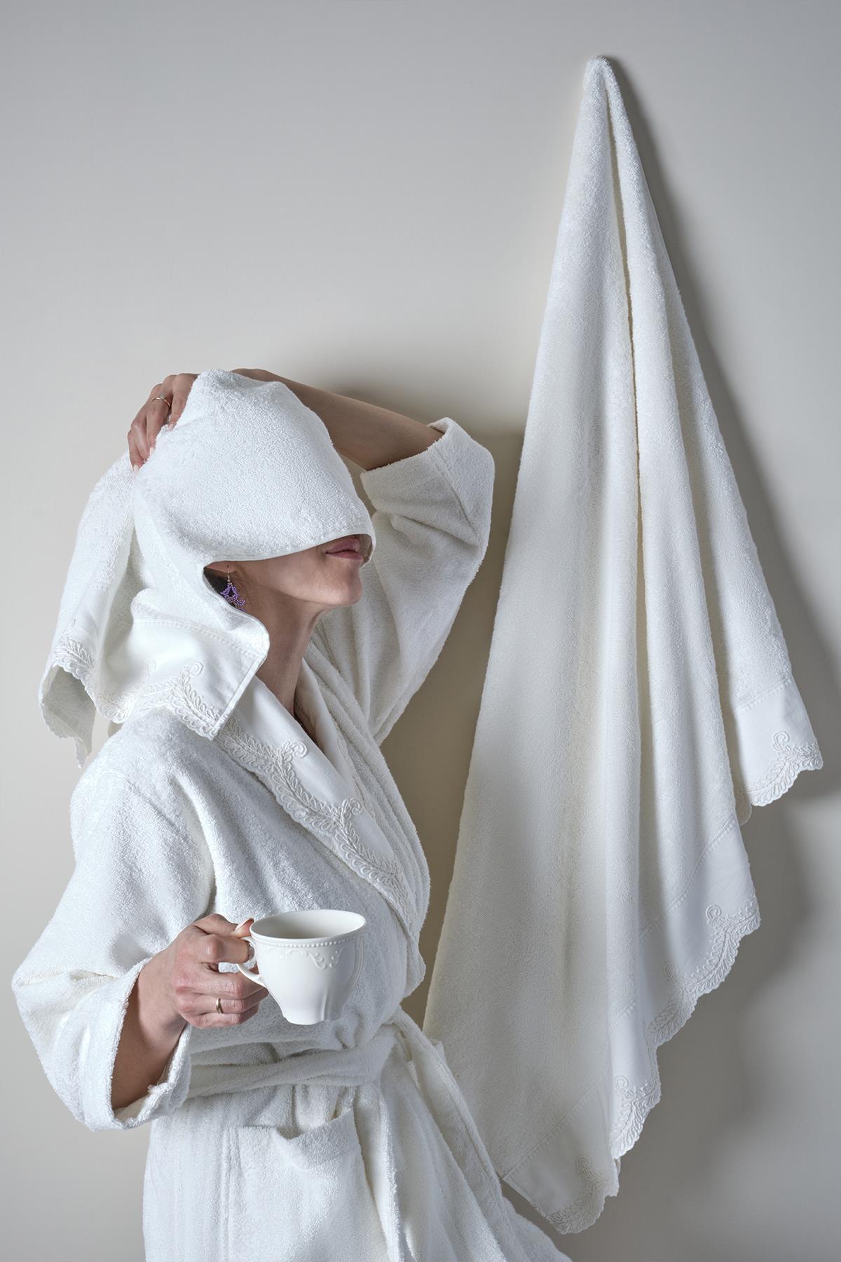 Femme en peignoir en bambou blanc tenant une tasse, avec une serviette suspendue au mur