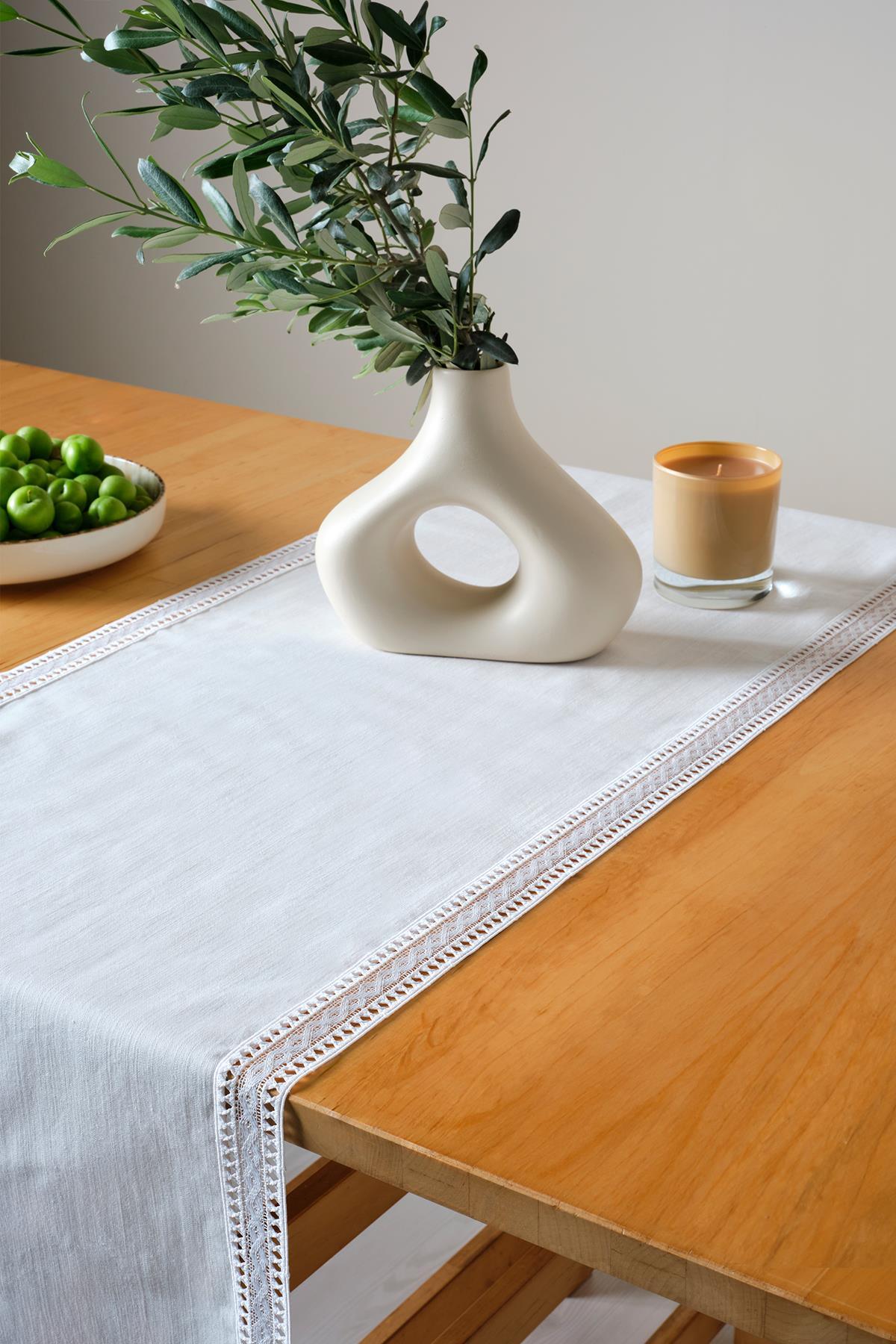 Chemin de table en dentelle blanche sur une table rectangulaire en bois, avec un vase moderne, des fruits verts et une décoration de bougies.